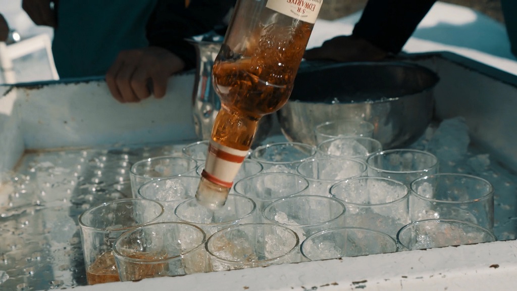 A close-up of a bottle of amber-colored liquor being poured into clear glasses filled with ice