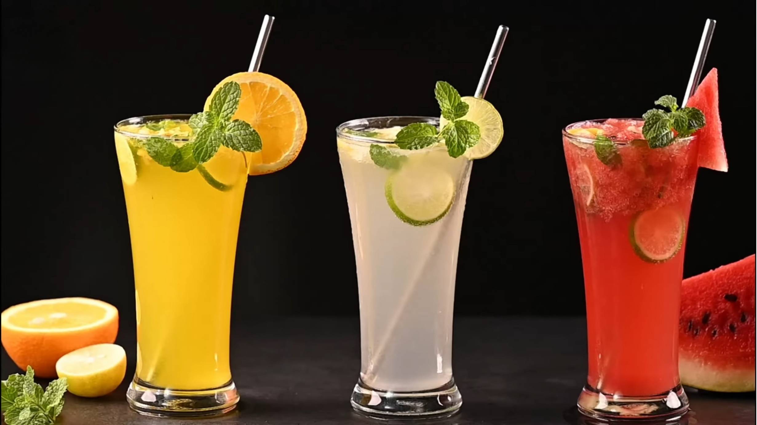 Three colorful Mocktails in tall glasses, garnished with mint, citrus slices, and watermelon