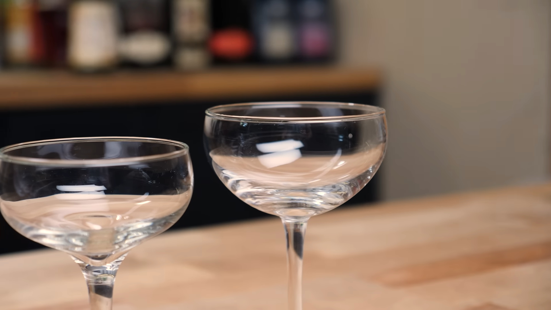 A close-up of two Nick and Nora glasses on a wooden surface with a blurred bar background