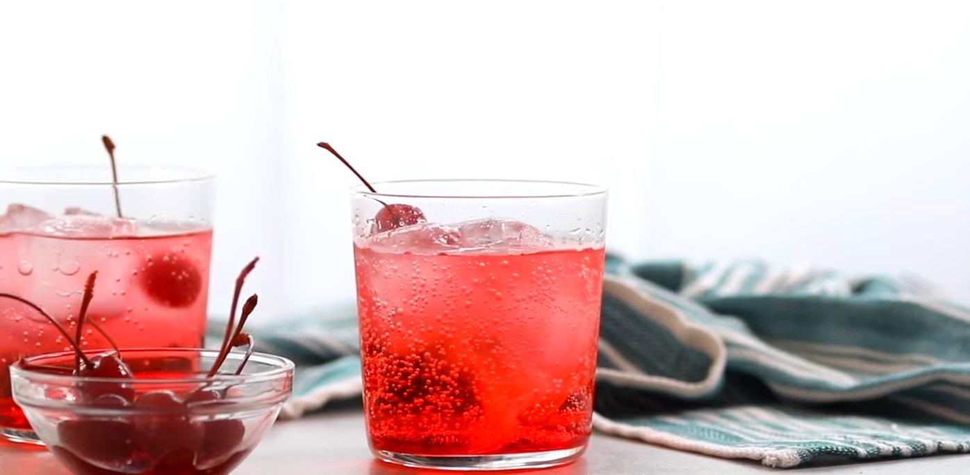 A bright red Shirley Temple Mocktail with ice, cherries, and bubbles, placed beside a bowl of maraschino cherries