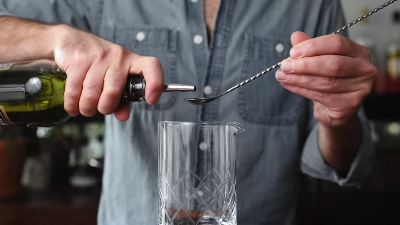 A Person Pours Liquor from A Bottle Into a Glass While Holding a Bar Spoon in The Other Hand