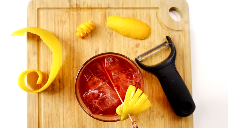 A Cocktail with A Lemon Peel Garnish Sits on A Wooden Cutting Board Next to A Peeler and Tweezers