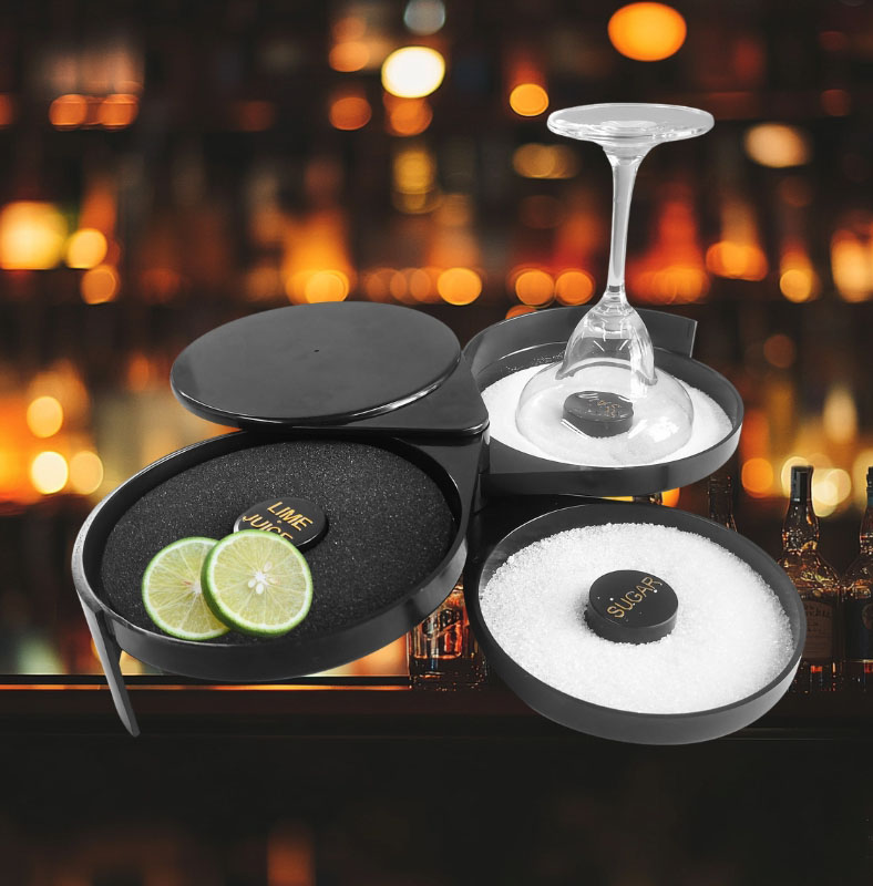 A three-tiered black cocktail rimmer set on a bar counter, containing lime juice, salt, and sugar, with a margarita glass being rimmed in salt