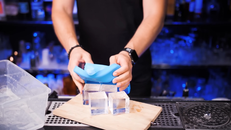 A Person Uses a Blue Ice Mold to Crush Large Ice Cubes on A Bar Counter