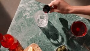 A Hand Pours a Measured Amount of Red Liquid Into a Glass with Ice on A Green Marble Countertop