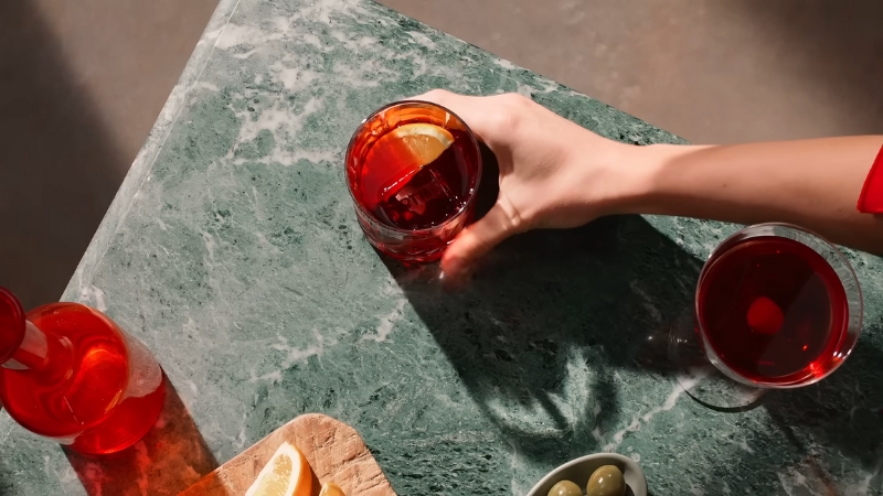 A Hand Reaches for A Negroni Cocktail Garnished with An Orange Slice on A Green Marble Table