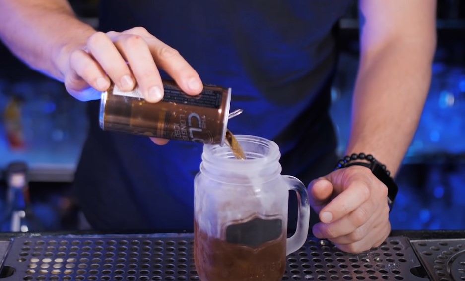 A bartender expertly pours a dark brown beverage into a frosty glass jar