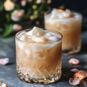 Close-up of two crystal glasses filled with creamy pineapple coconut cocktails over ice, surrounded by soft pink flower petals on a textured surface