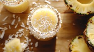 Close-up of a cocktail glass rimmed with toasted coconut flakes, filled with a creamy pineapple coconut cocktail. Slices of fresh pineapple and scattered coconut flakes on a wooden surface